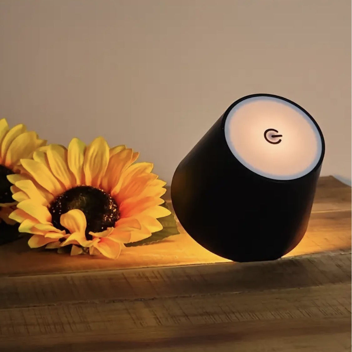 Black cylindrical lamp with a glowing light bulb on a wooden surface next to sunflowers.