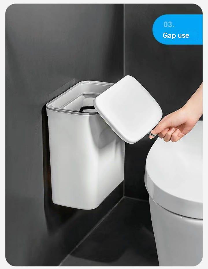 White trash bin with lid attached to a wall next to a toilet.